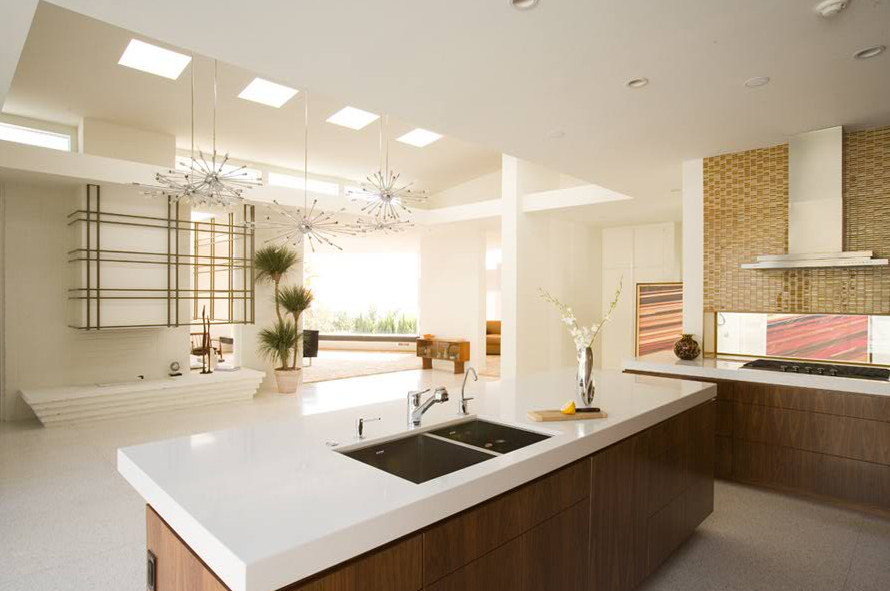 Modern kitchen with large white island featuring double undermount sinks, brown wood cabinetry, and a cooktop with a stainless steel hood, a