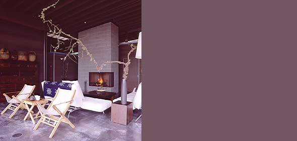 Interior view of a living room with a concrete fireplace, white seating, wooden folding chairs, and decorative branches in a vase.