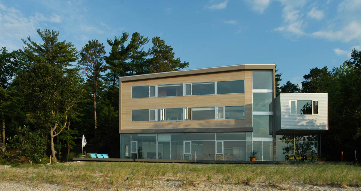 Modern three-story house with horizontal wooden and light siding, large glass windows, and a flat roof, situated by a sandy and grassy area.