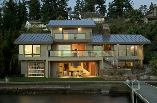 Modern two-story lakeside house with large windows and metal roofs, viewed at dusk with interior lights on.
