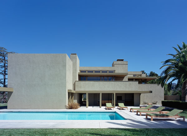 View of a modern beige stucco two-story house with rectangular shapes and a swimming pool in front under a clear blue sky.