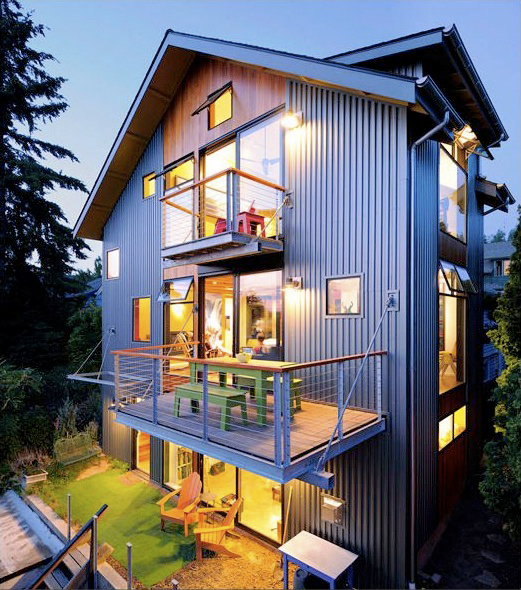 Night view of a modern three-story house with metal siding, wooden accents, and two balconies with outdoor seating.