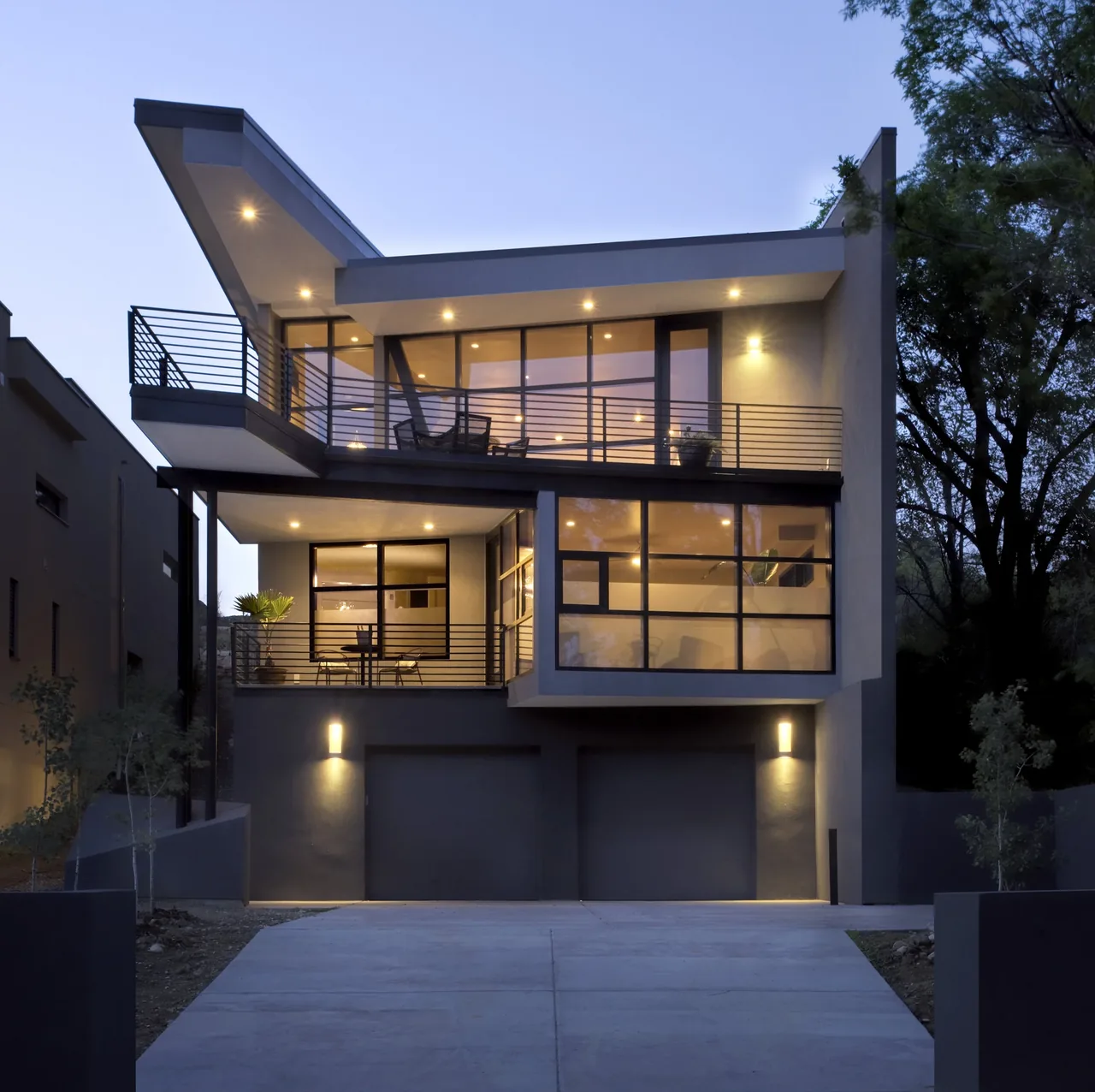 Front exterior view of a modern three-level house with large windows, balconies, and garage doors, illuminated at dusk.