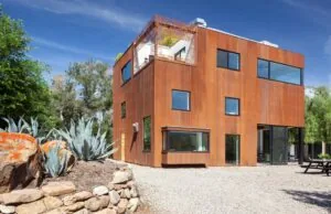 Three-story modern residence with rust-colored exterior cladding surrounded by gravel and desert landscaping under a clear blue sky.