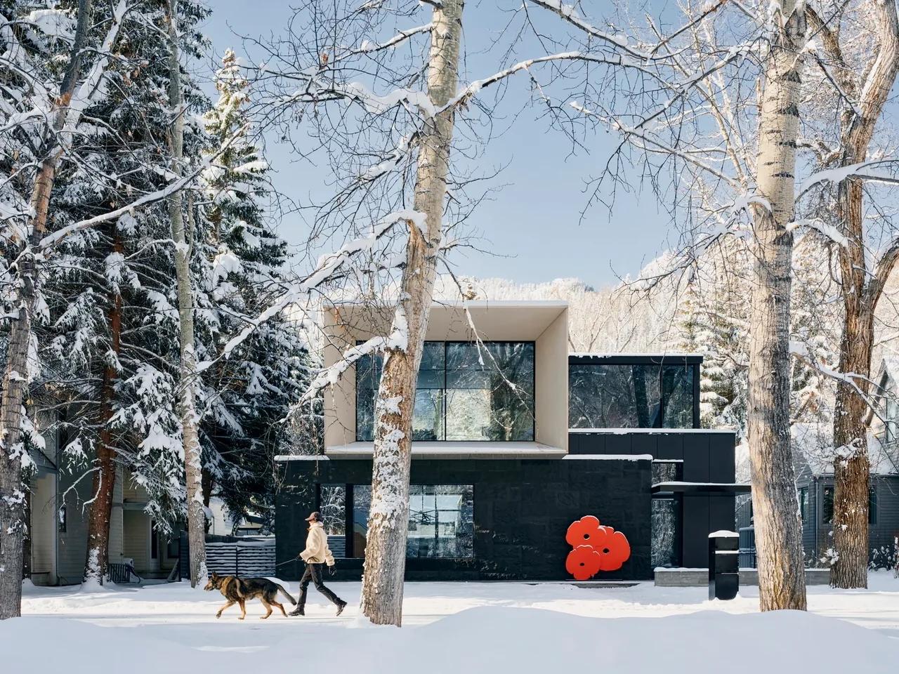 Modern two-story house with large windows and black lower facade, surrounded by snow-covered trees and snow on ground, with a person walking