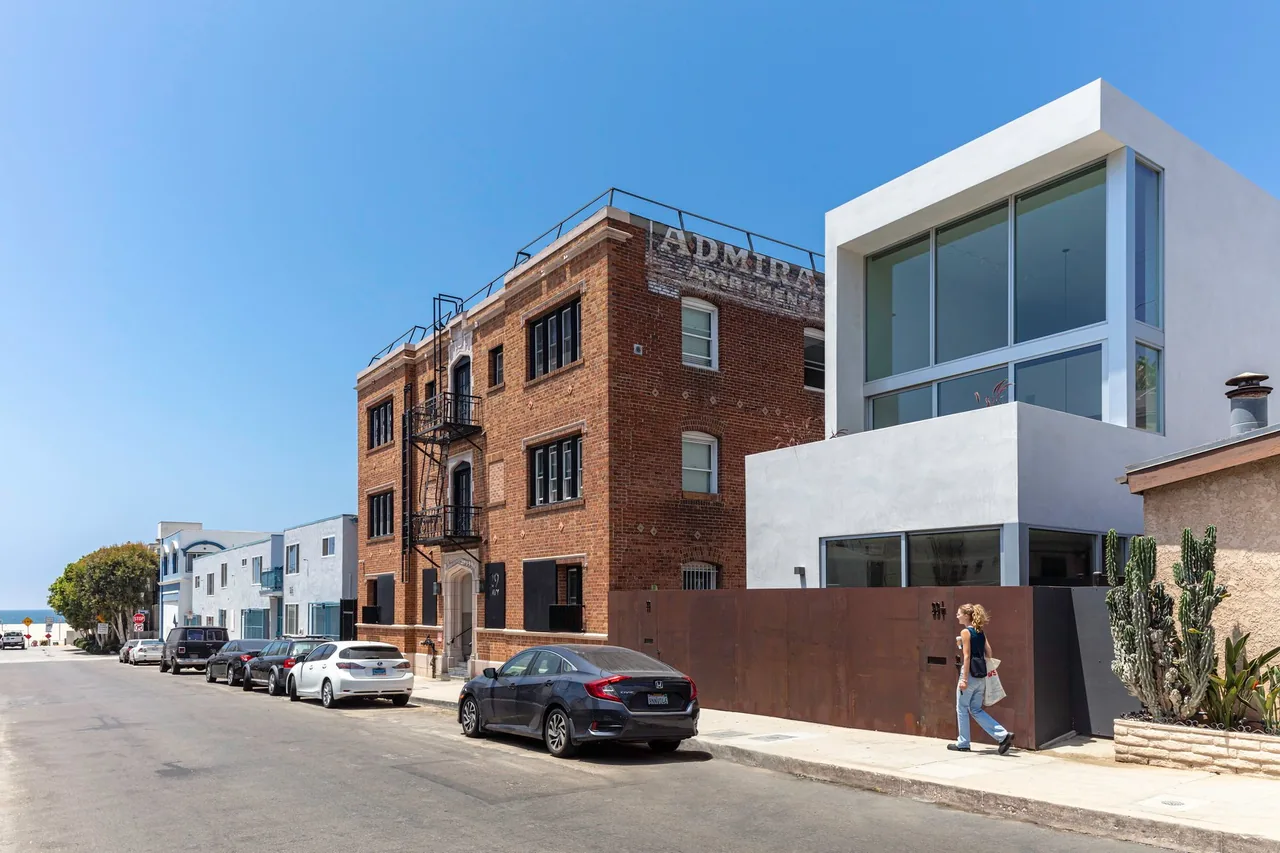 Street view showing a modern white two-story building with large windows next to a historic red brick three-story apartment building with a