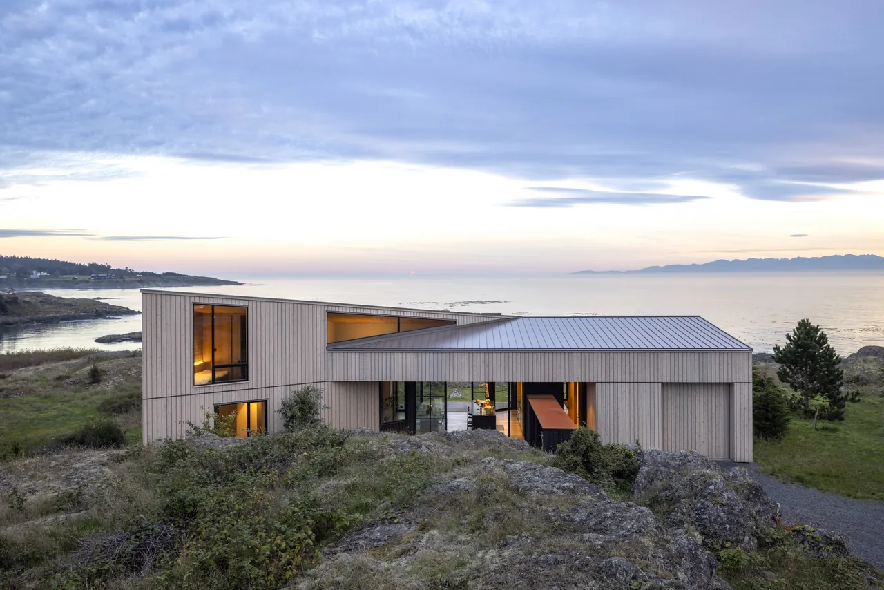 Modern wood-clad house overlooking a rocky shoreline and the sea at dusk.