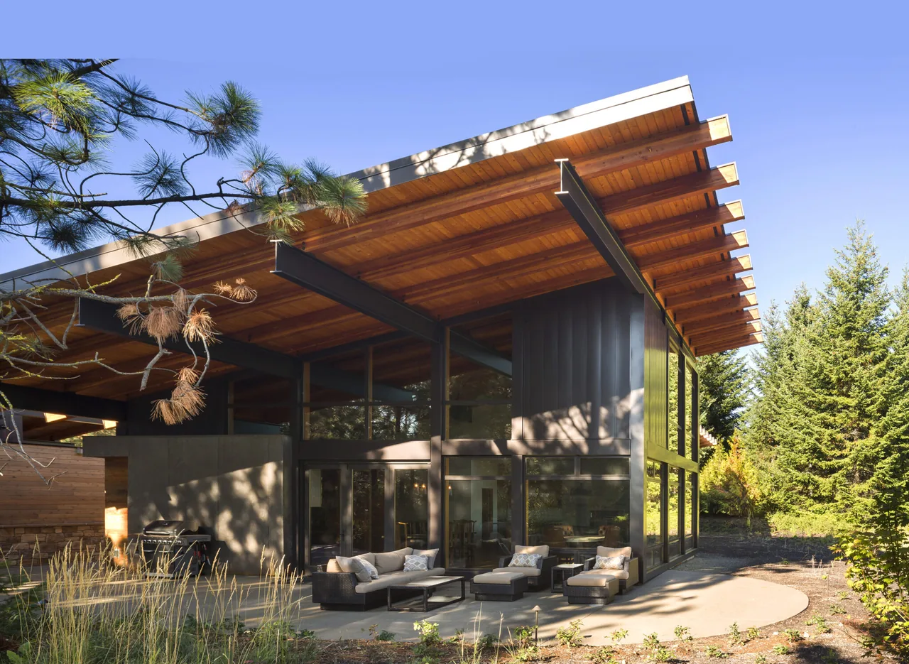 Modern house exterior with sloped wood-paneled roof, black steel beams, and large floor-to-ceiling windows opening onto an outdoor patio set
