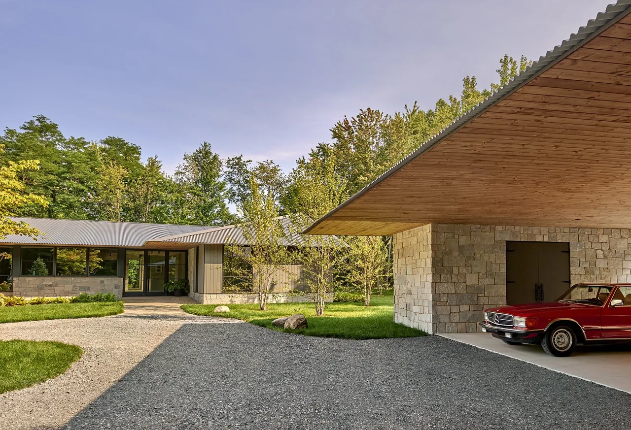 Exterior view of a single-story modern home with stone and wood materials, featuring a driveway leading to a carport with a red vintage car,