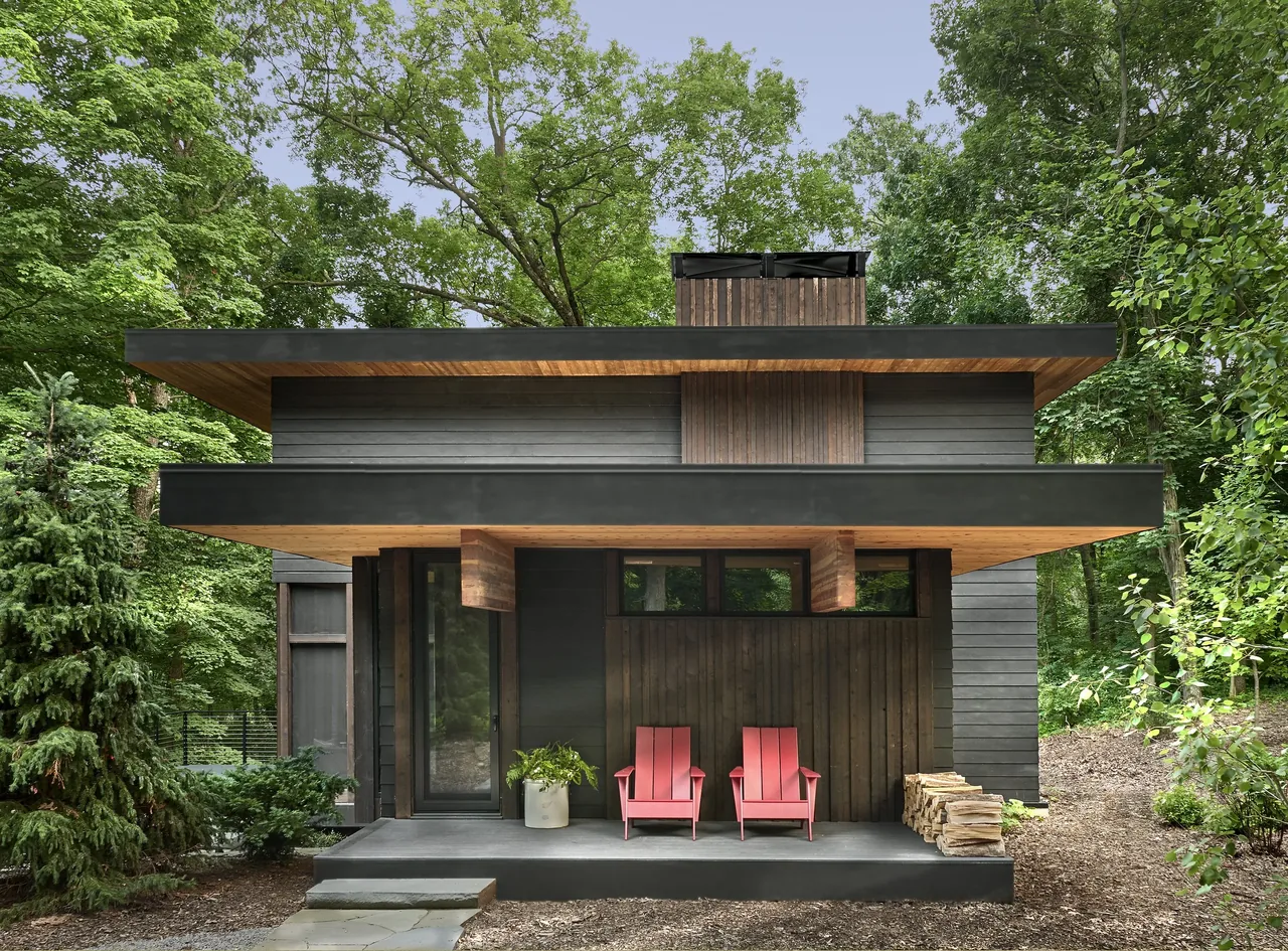 Front view of a modern two-story retreat house with dark horizontal siding and deep overhanging flat roof eaves, flanked by dense trees.