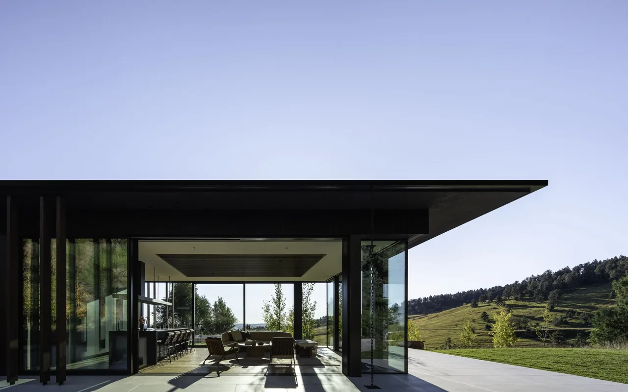 Modern house exterior with large floor-to-ceiling glass walls and a flat roof, opening to a patio with views of rolling hills and trees.