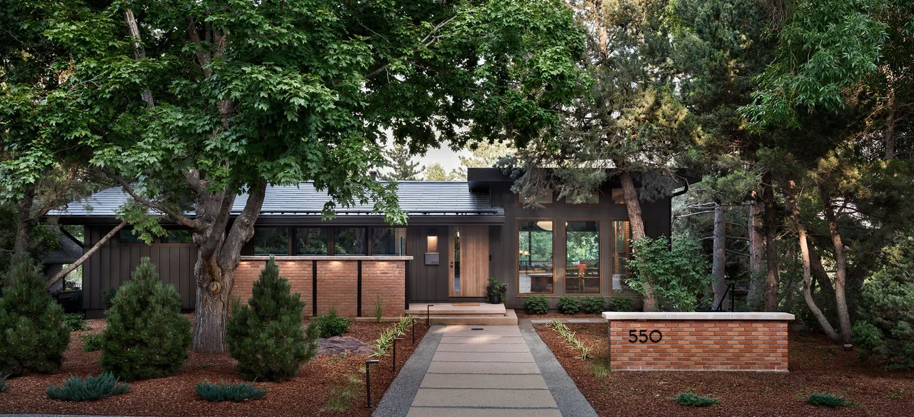 Exterior front view of a single-story residence with dark vertical siding, brick planters, and large windows, surrounded by mature trees and