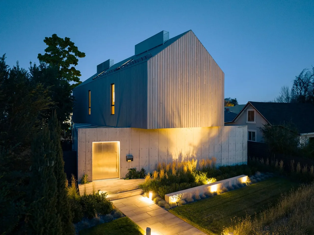 Evening view of a modern two-story house with vertical wood siding upper level and concrete lower level, featuring illuminated entrance and庭