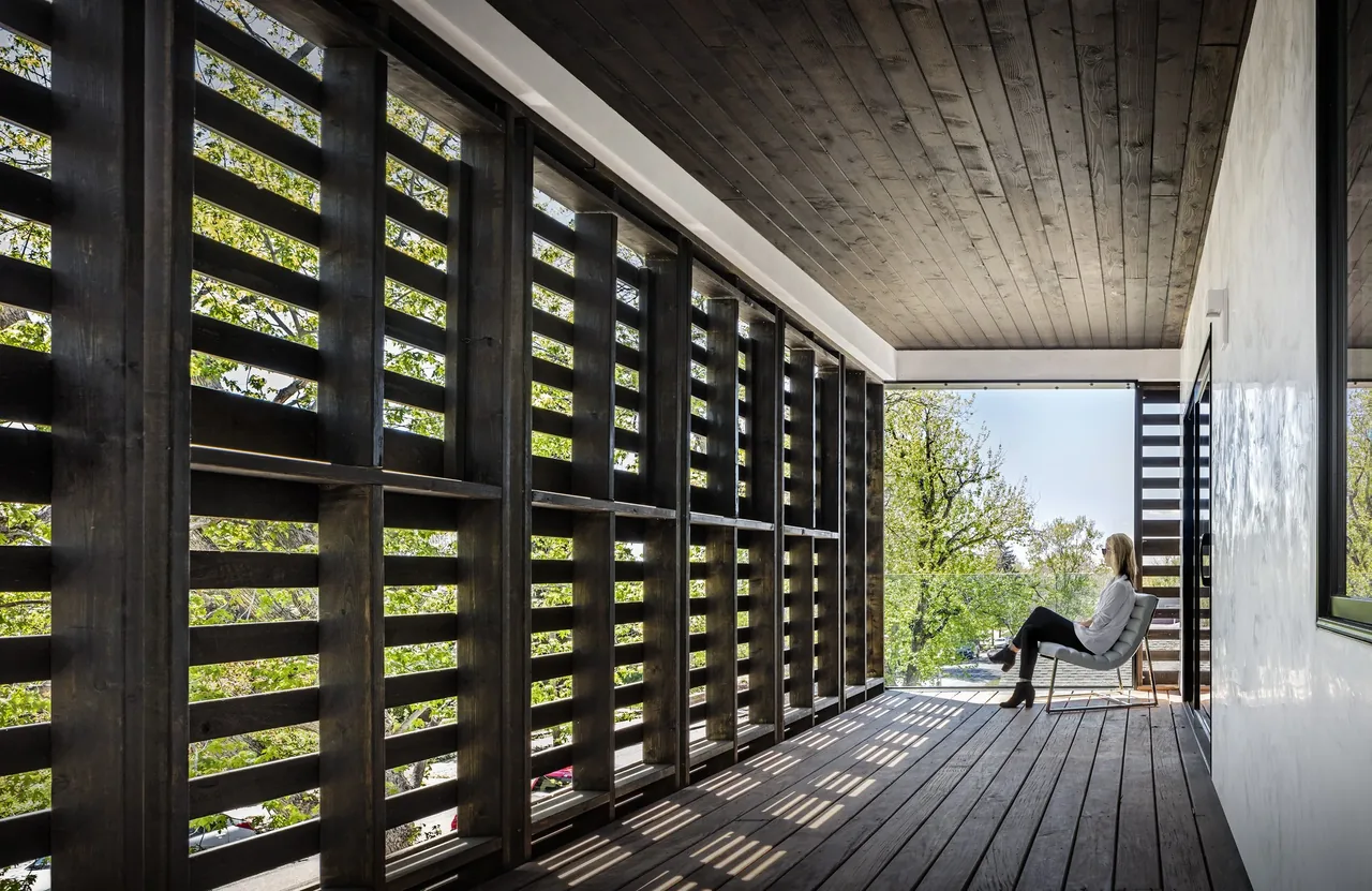 View of a covered patio with wooden slatted walls and ceiling, featuring a seated woman looking outside at trees under daylight.