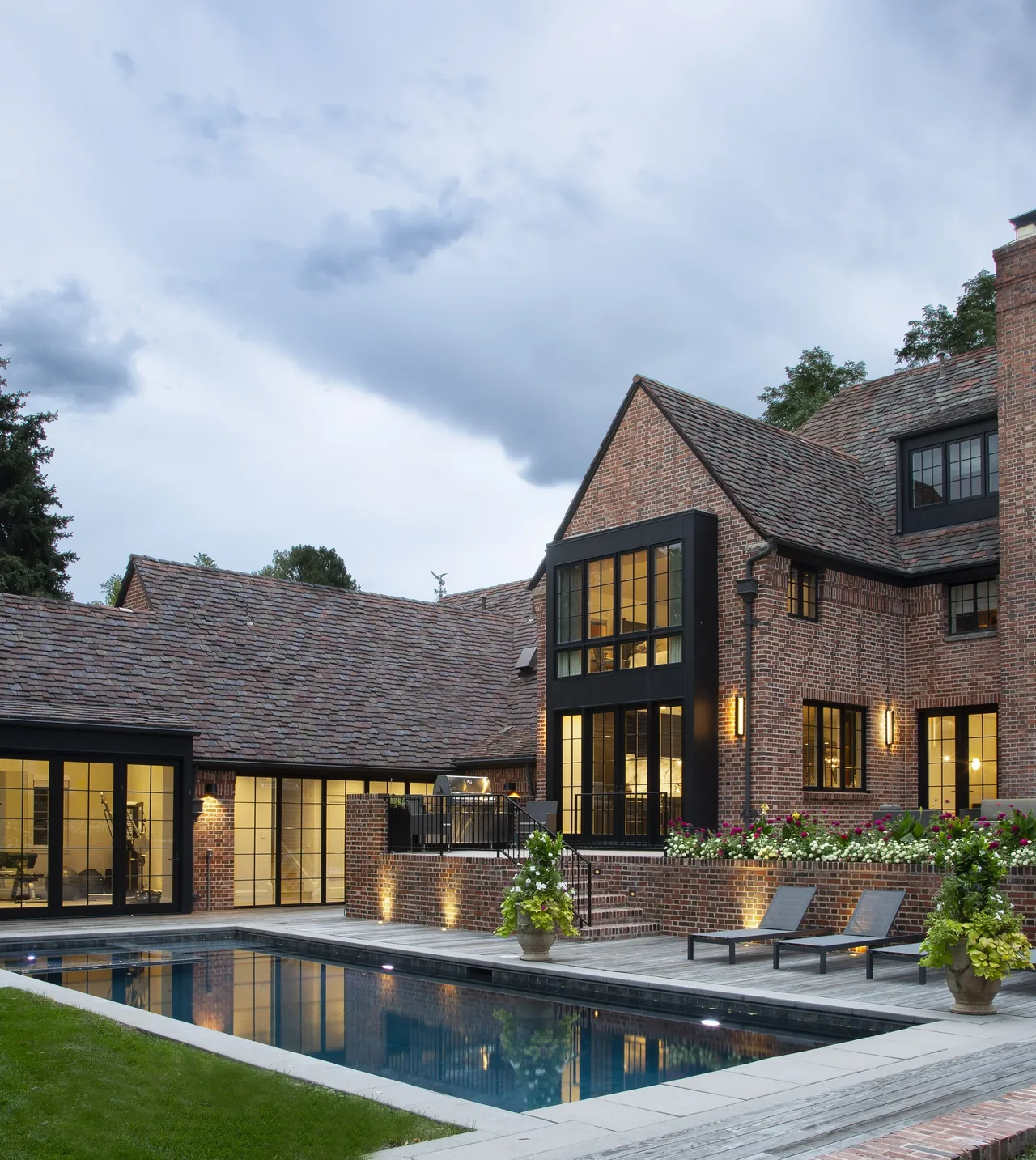 Exterior view of a brick house with multi-pane black window frames and a pool in the foreground at dusk.