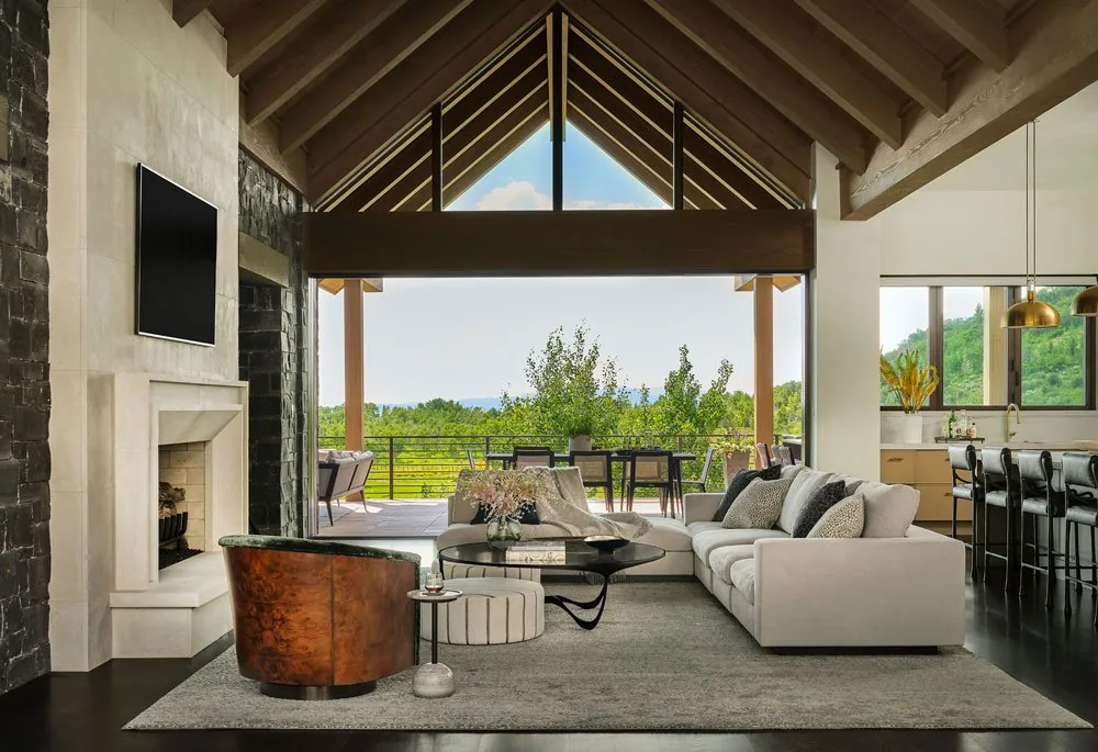 Living room with vaulted wood ceiling, stone fireplace, and large sliding glass doors opening to a patio with outdoor seating and greenery.