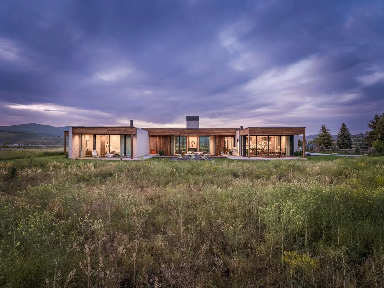 Modern single-story house with wood and stucco facade set in a natural grassy landscape under cloudy sky at dusk.