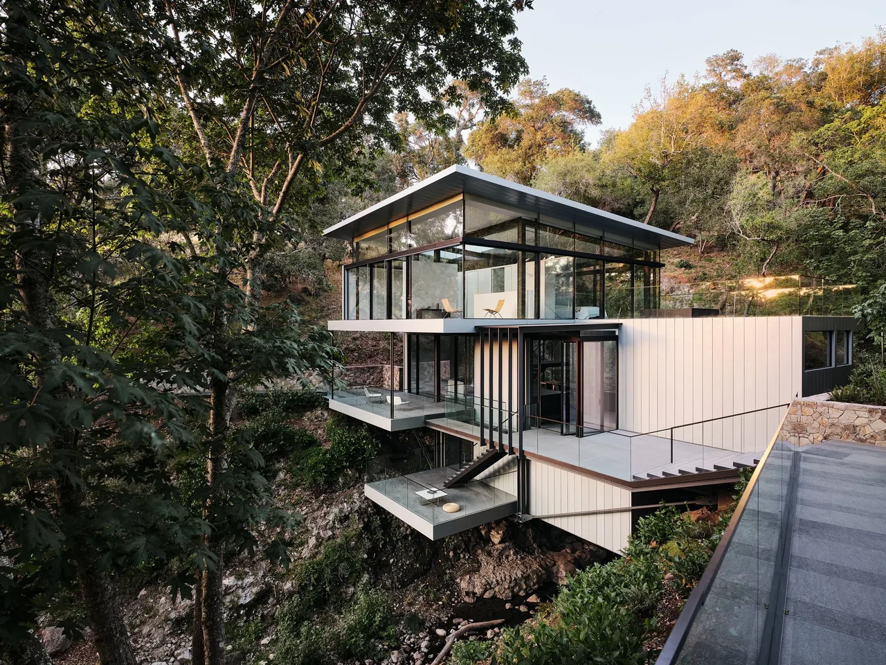 Modern house suspended over a rocky stream, surrounded by trees and forest landscape, with two cantilevered terraces and glass walls under a