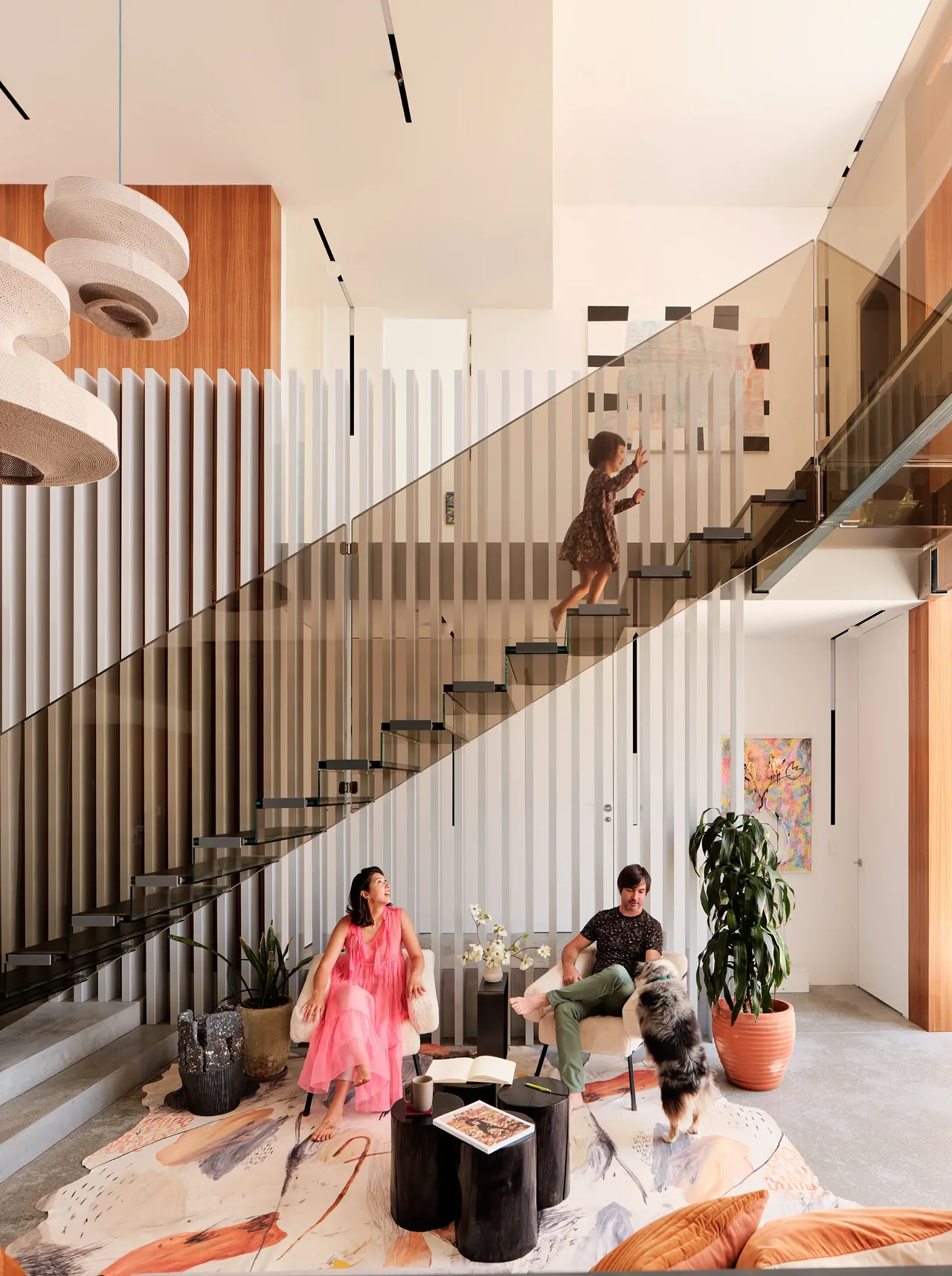 Modern living room with floating glass stairs and vertical slats, showing a woman sitting, a man interacting with a dog, and a child walking