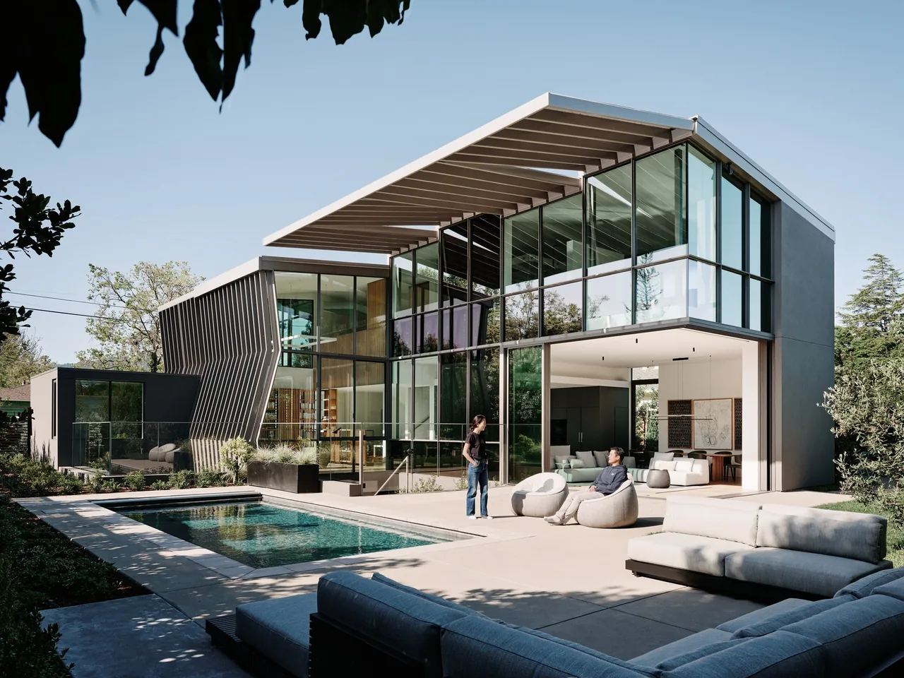 Modern two-story house with extensive floor-to-ceiling glass, angled metal panels, and a flat roof with overhang, showing an open patio and
