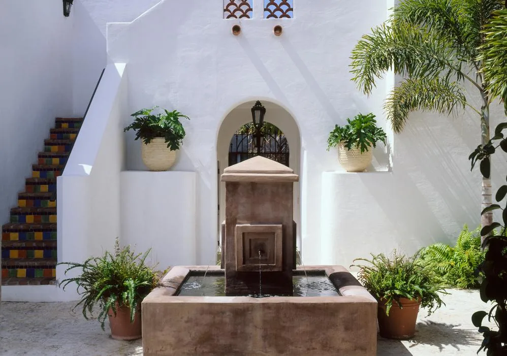 Outdoor courtyard with white stucco walls, a central stone water fountain, potted plants, and colorful tiled stairs.