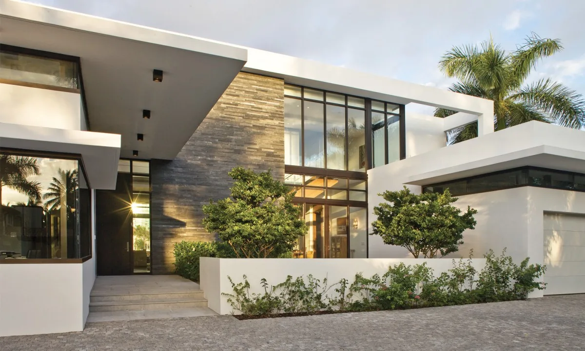 Modern residence exterior featuring white stucco walls, large floor-to-ceiling windows with dark frames, and a stone accent wall under a sl3