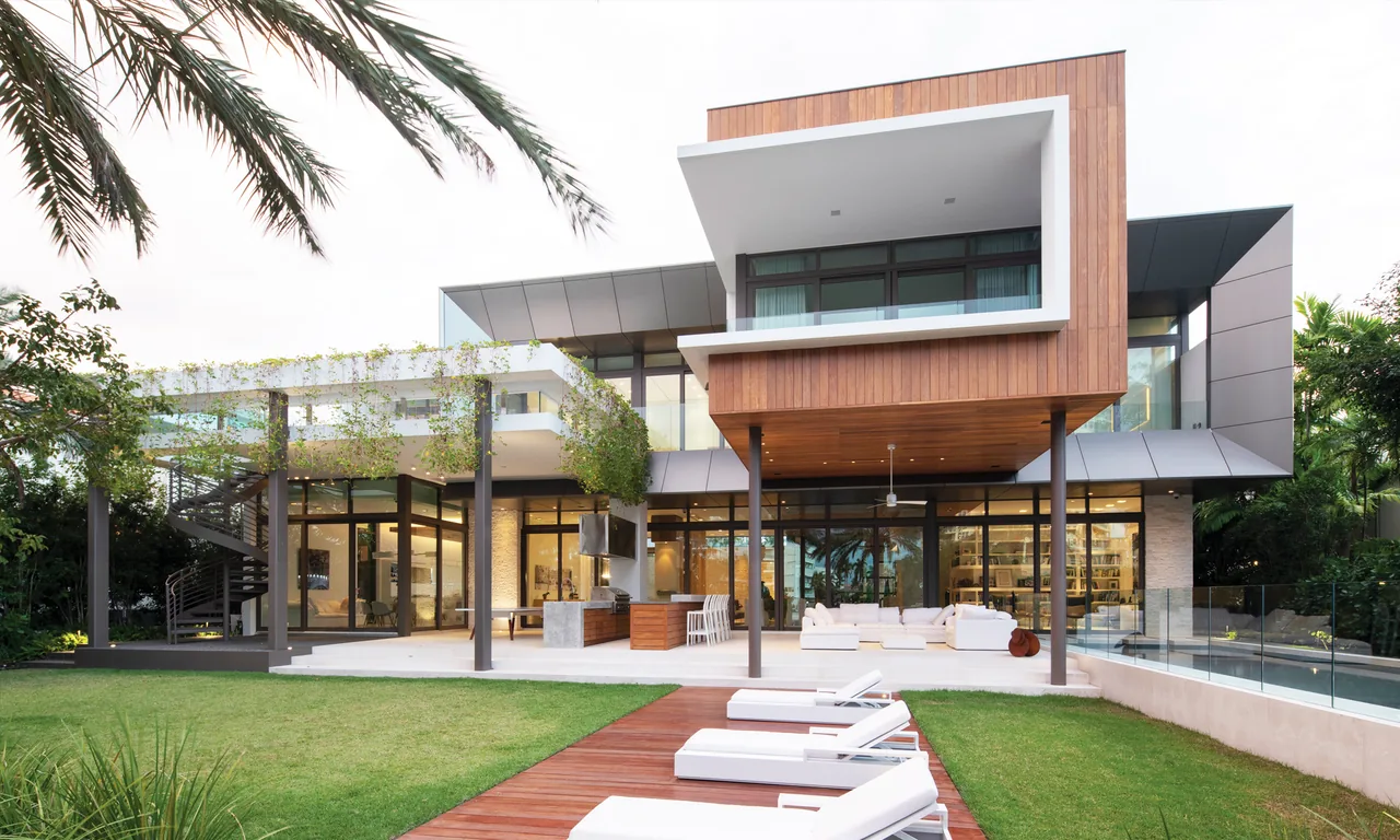 Modern two-story house with wood and metal panel exterior, large glass walls, outdoor lounge area, and poolside lounge chairs on a wooden on