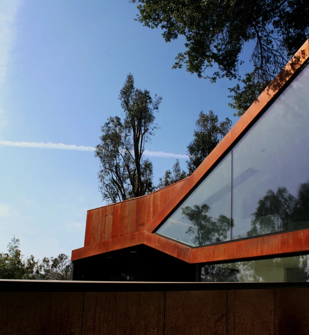 Angular view of a modern building exterior with rust-colored metal cladding and large slanted floor-to-ceiling glass windows, trees and blue