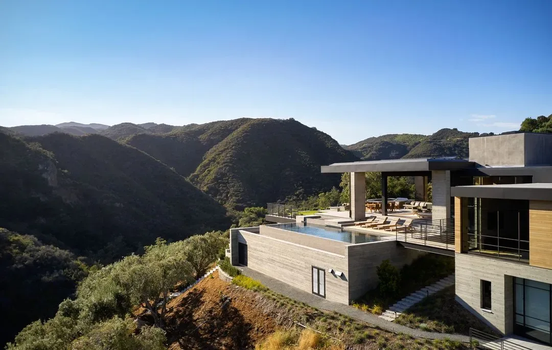 Modern hillside residence with a rectangular pool and covered patio overlooking forested mountains under a clear sky.