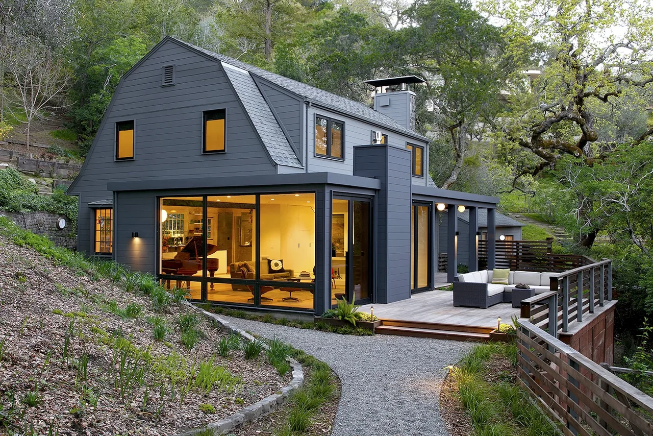 Dark gray modern barn-style house with large windows and a lighted interior, surrounded by greenery and a wooden deck.