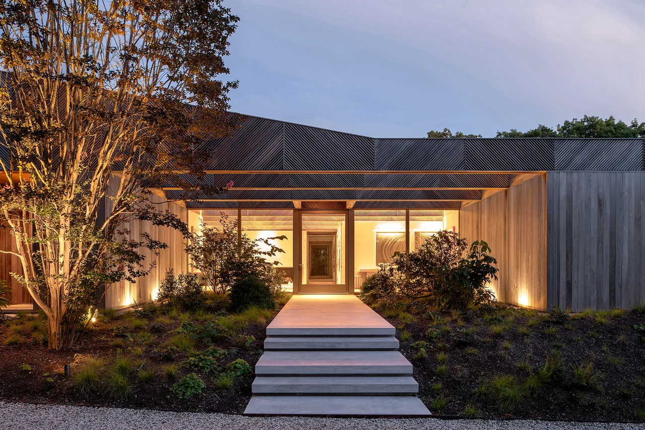 Exterior front view of a modern house at dusk with wood siding, a central glass entry door, and softly lit landscaping with steps leading to