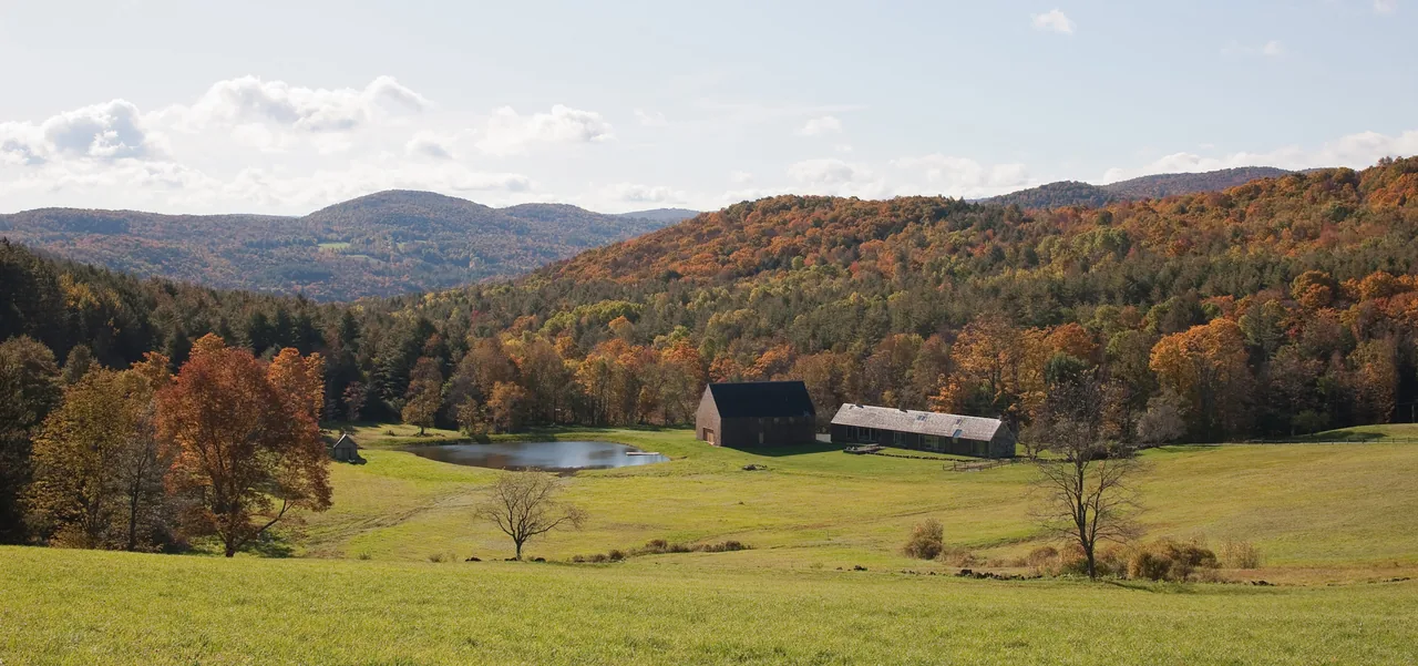 Two farm buildings near a pond in a green field surrounded by autumn forest and hills under a partly cloudy sky.