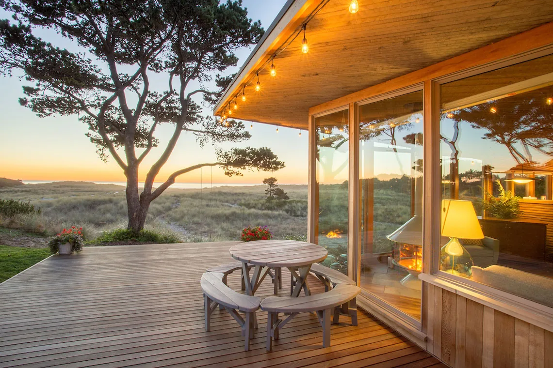 Wooden deck at sunset with round picnic table, curved benches, and reflections of a living room with fire and lamp in large glass windows.