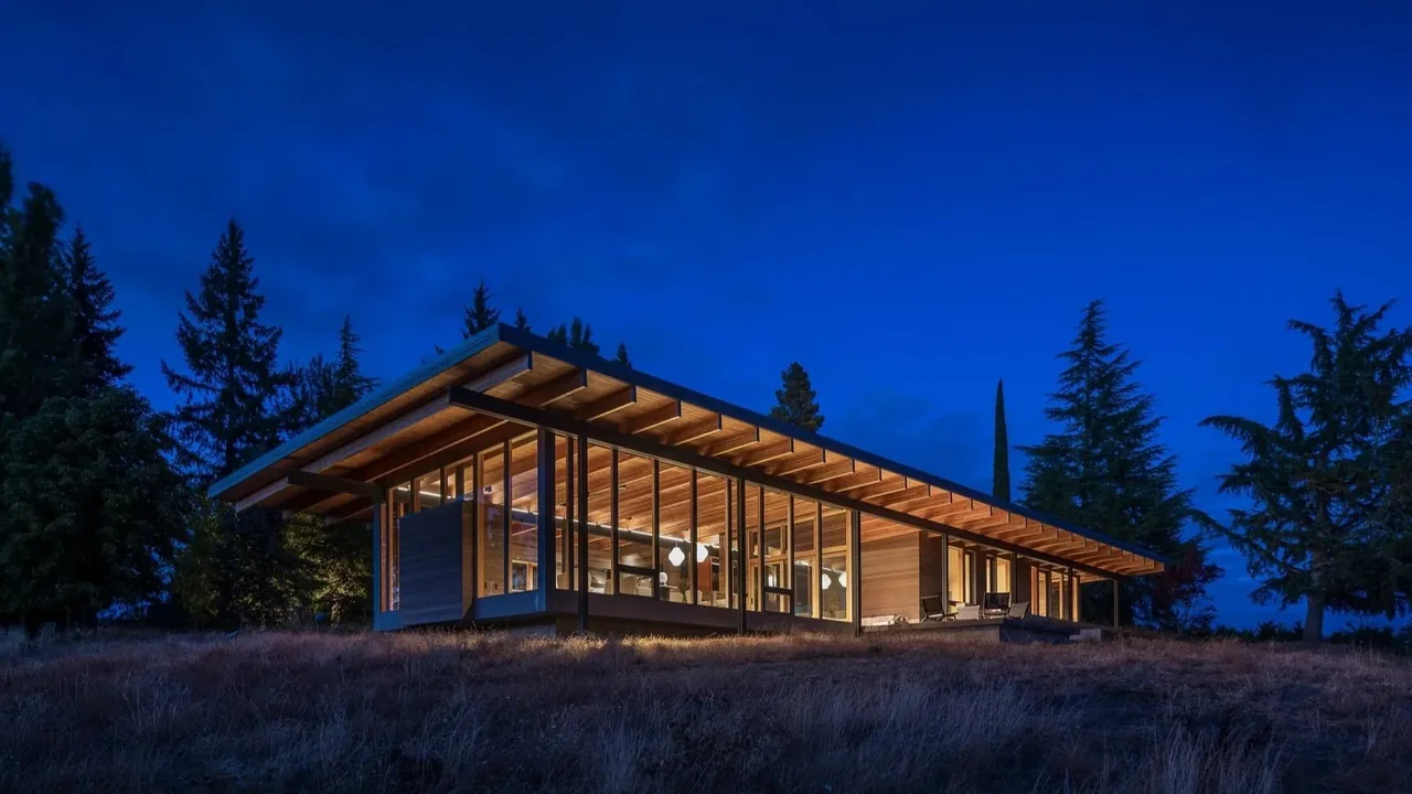 Night view of a modern one-story house with extensive floor-to-ceiling glass walls and a wood overhanging flat roof.
