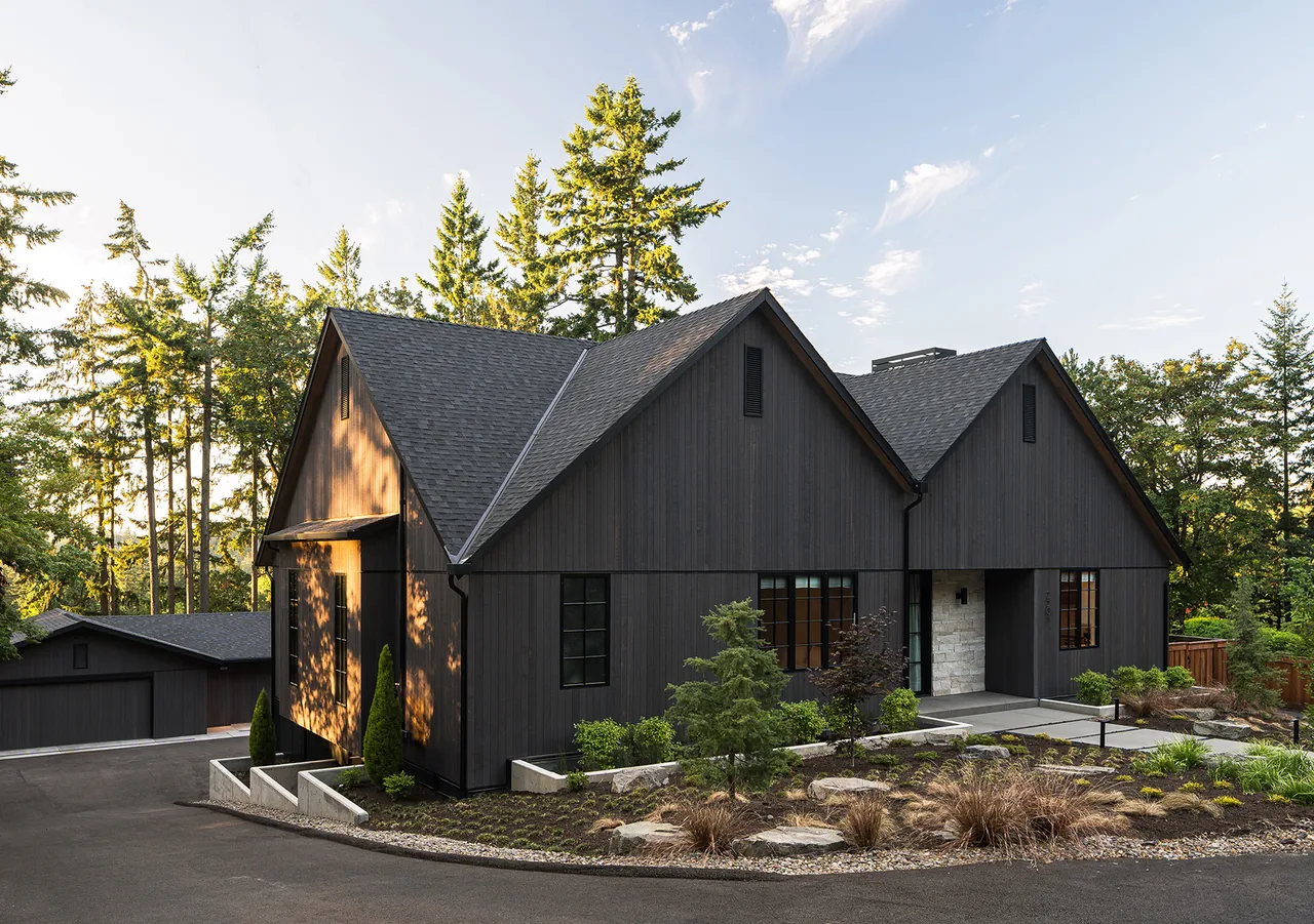 Black wooden house with gable roofs surrounded by trees under a partly cloudy sky.