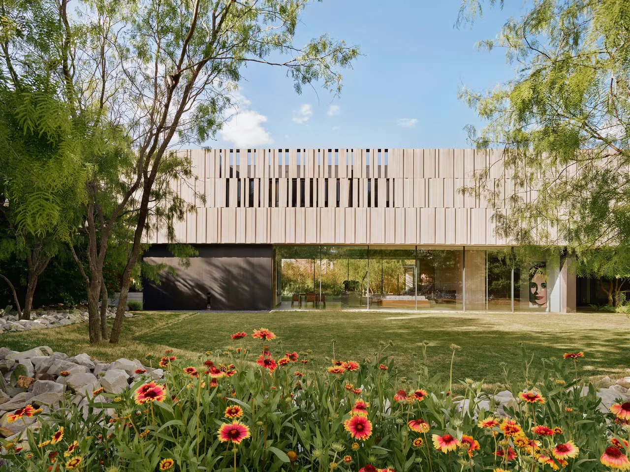 Modern two-story house with upper facade of vertical wood panels and large glass windows on ground level, set in a green garden with trees.