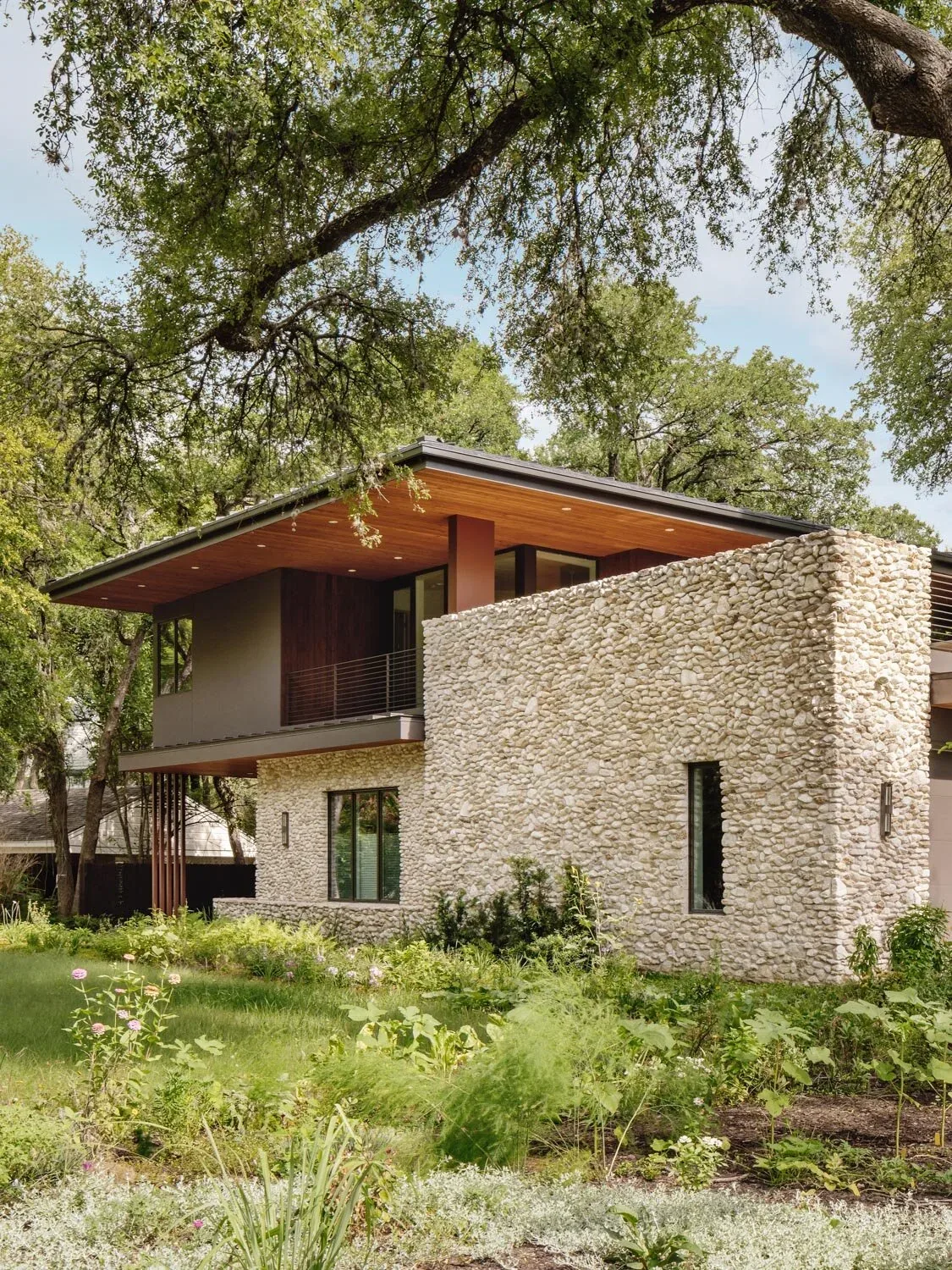 Two-story modern house with a stone exterior wall, large windows, and a wide flat roof surrounded by trees.
