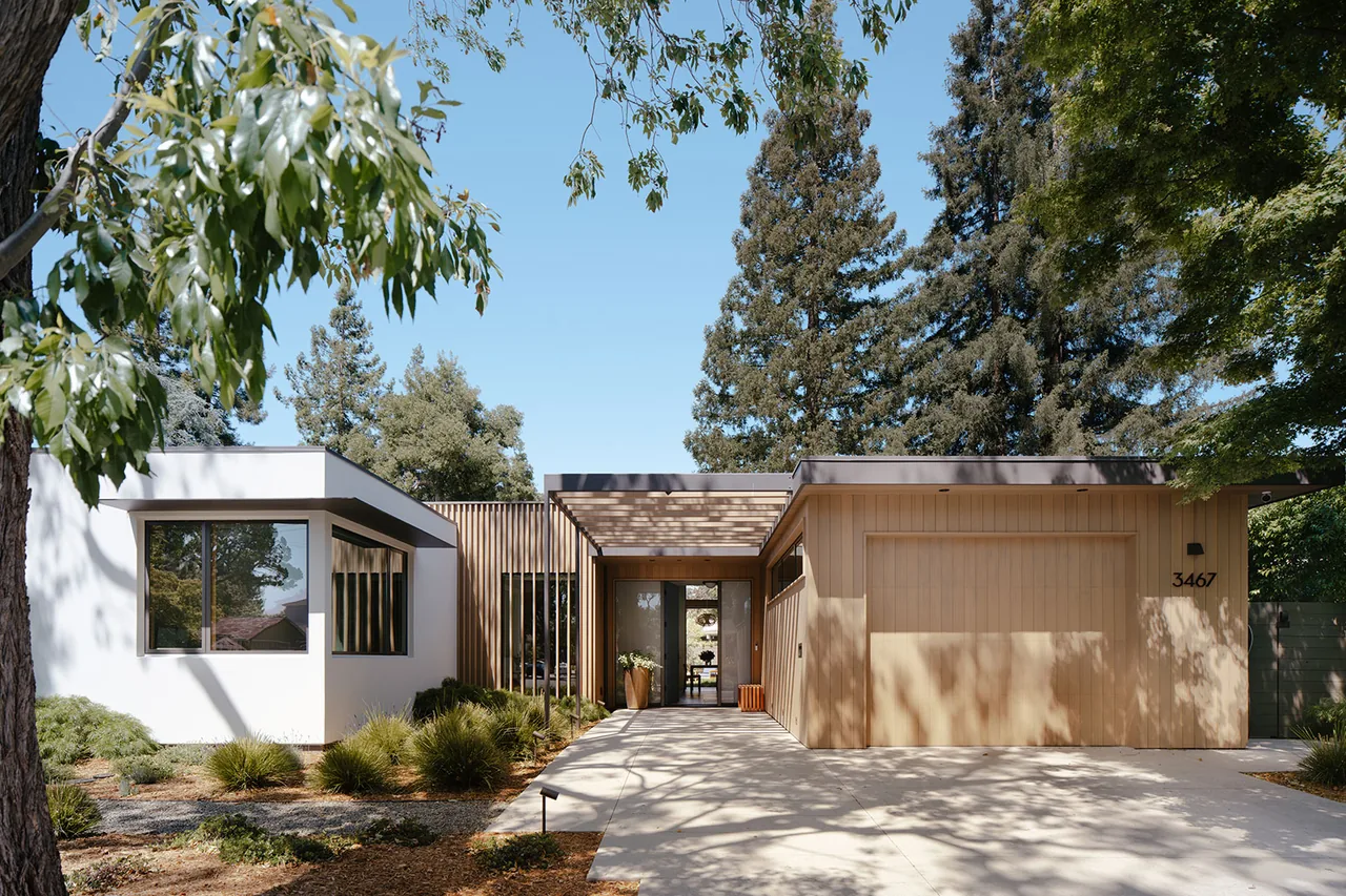 Modern single-story home with a flat roof, wooden garage door, and white stucco exterior walls, surrounded by trees.