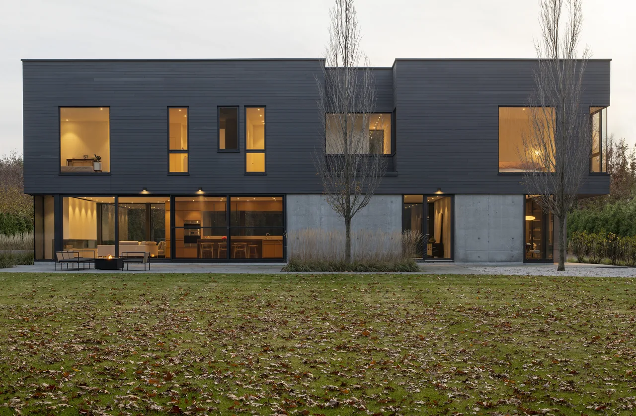 Exterior rear view of a two-story modern house with dark wood siding and large windows at dusk.