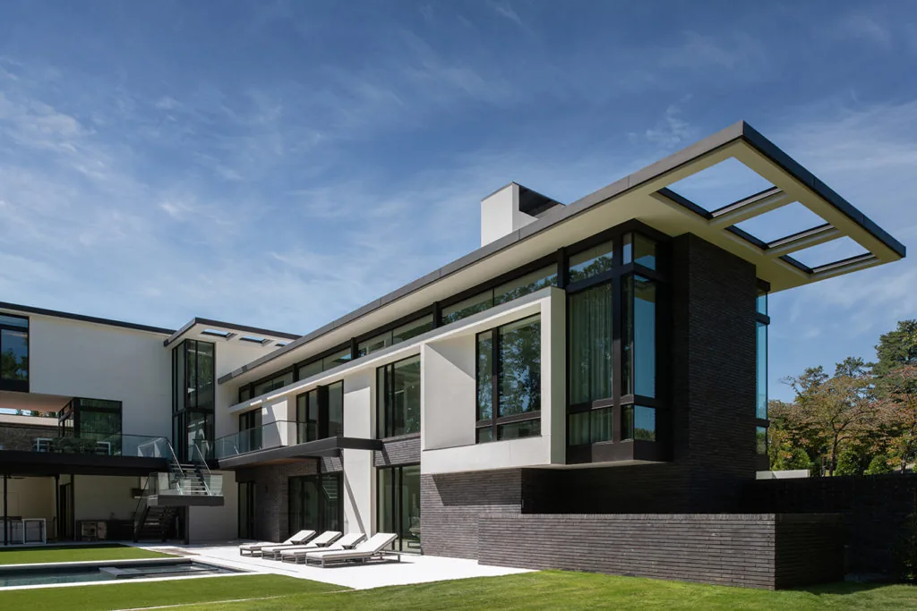 Modern two-story house with large black-framed windows and a cantilevered roof section over a patio with lounge chairs.