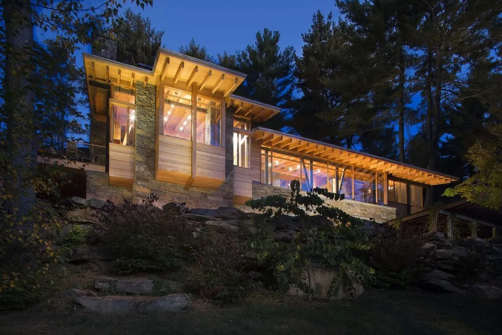 Evening view of a modern house with large windows and wooden eaves surrounded by trees.