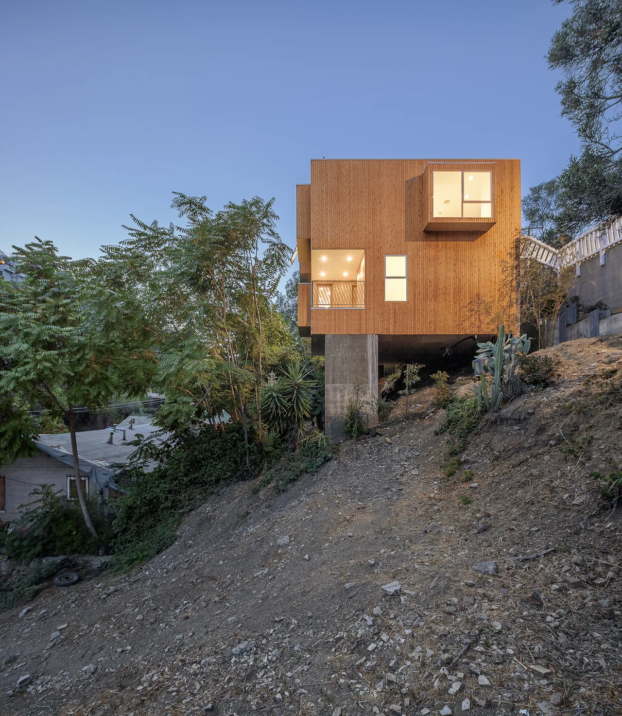 Wood-clad house elevated on concrete pillars on a steep hillside with surrounding vegetation under a clear sky.