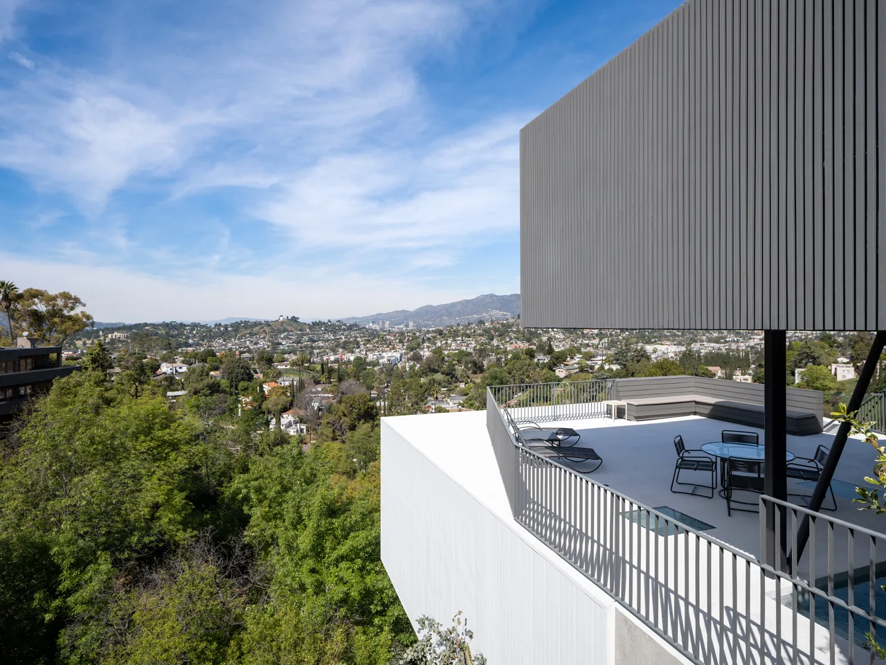 Modern house balcony with metal railing and outdoor seating overlooking a cityscape with hills and trees.