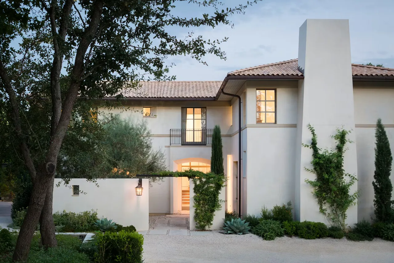 Exterior view of a two-story house with light stucco walls, tile roof, climbing vines, and a rustic lantern hanging on a metal trellis at an