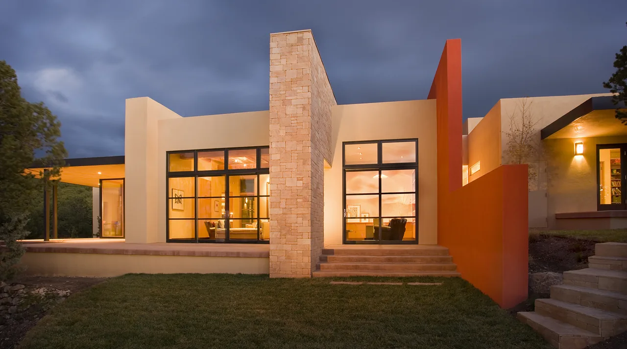 Modern flat roof house exterior with large black-framed windows and stone chimney at dusk.