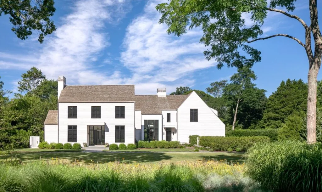 White two-story house with black windows and a gable roof surrounded by greenery under a partly cloudy blue sky.