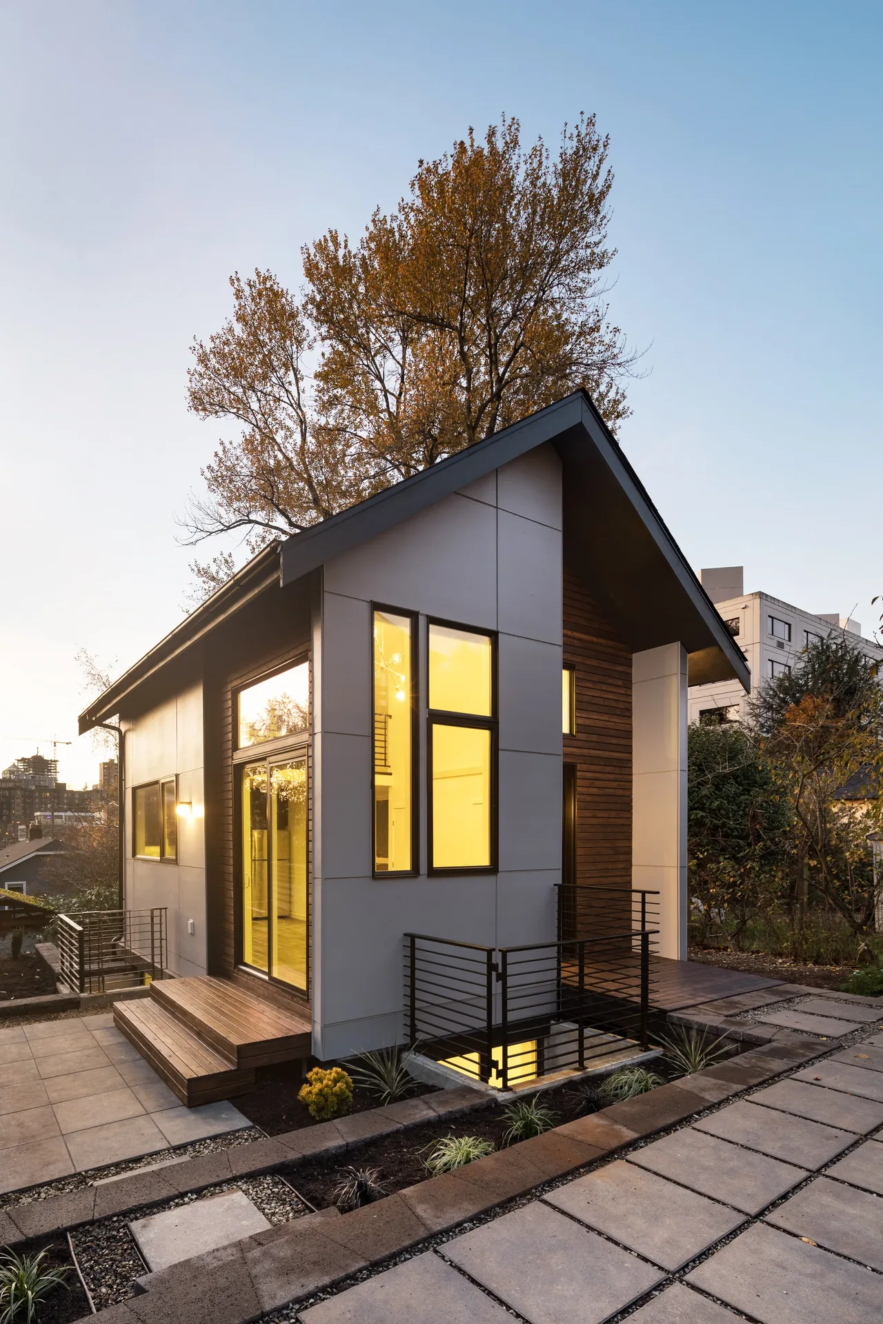 A small modern detached dwelling with wood siding and large windows in an urban garden area at dusk.