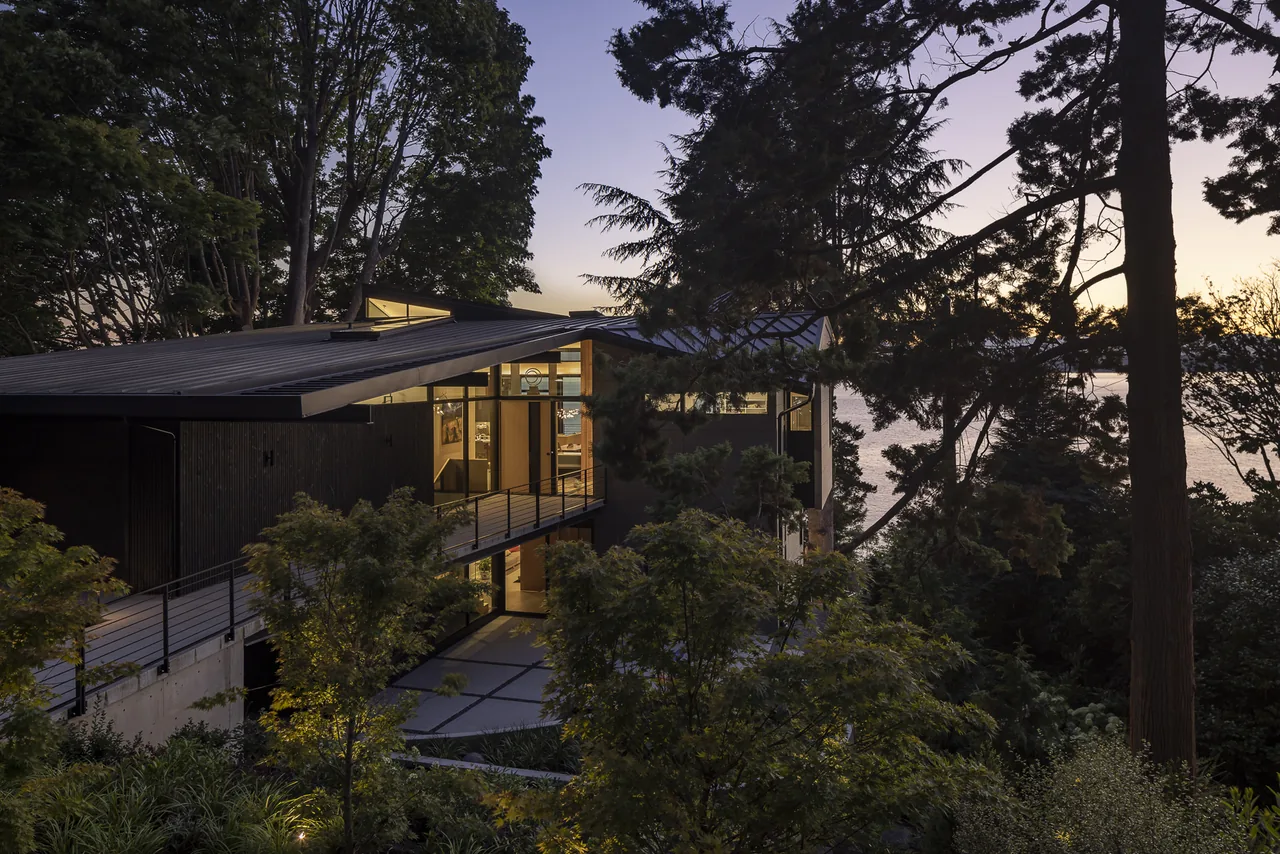 Modern home with flat roof and glass walls nestled among tall trees near a body of water at dusk.