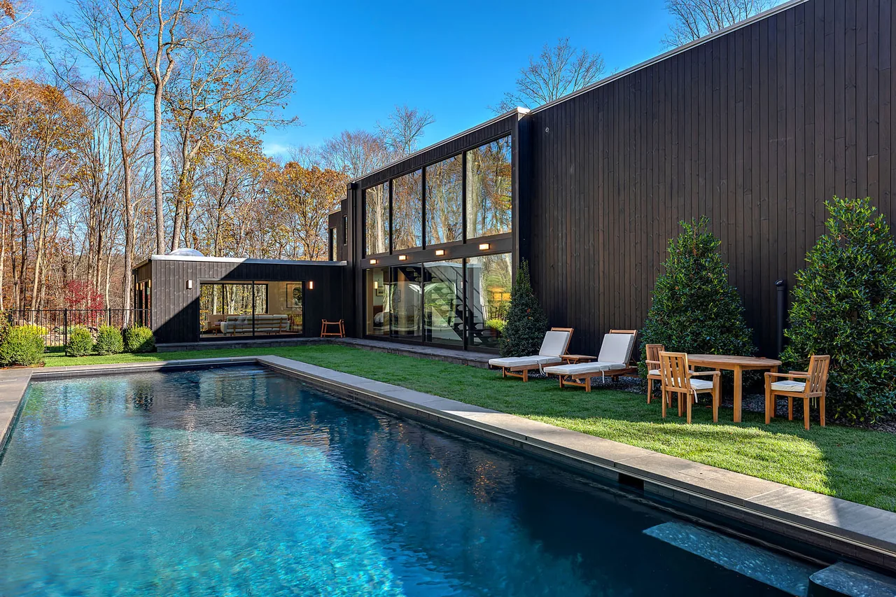 Dark wood-paneled modern house exterior with large glass windows and an adjacent swimming pool under a clear sky.