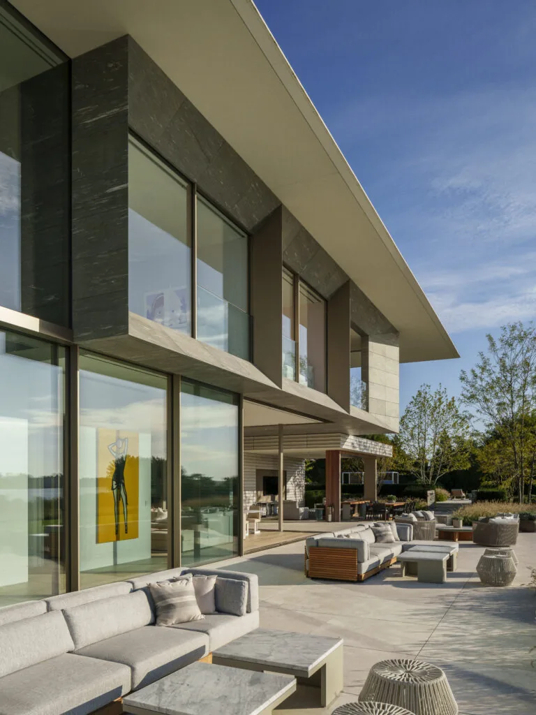 Modern two-story house with large glass windows and outdoor seating area on a spacious patio under a clear sky.